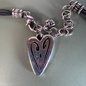 May include: A silver-toned heart pendant featuring an engraved abstract design, suspended from a silver-toned chain link connector and a black cord. The pendant includes a small heart-shaped charm near the clasp, which displays the letters EB.