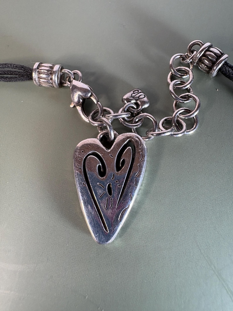 May include: A silver-toned heart pendant featuring an engraved abstract design, suspended from a silver-toned chain link connector and a black cord. The pendant includes a small heart-shaped charm near the clasp, which displays the letters EB.