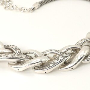 May include: Silver-toned necklace featuring a chunky chain design. The necklace has a textured, woven chain and a series of large, ornate links with etched detailing. The clasp is a simple lobster claw style.