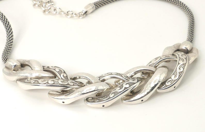 May include: Silver-toned necklace featuring a chunky chain design. The necklace has a textured, woven chain and a series of large, ornate links with etched detailing. The clasp is a simple lobster claw style.