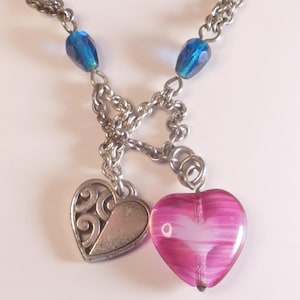 Puede incluir: Un collar de cadena de metal plateado con dos dijes en forma de coraz&oacute;n. Un dije es un coraz&oacute;n plateado s&oacute;lido con un patr&oacute;n de remolinos en relieve, y el otro es un coraz&oacute;n de vidrio rosa transl&uacute;cido con rayas blancas. Dos cuentas azules est&aacute;n unidas.