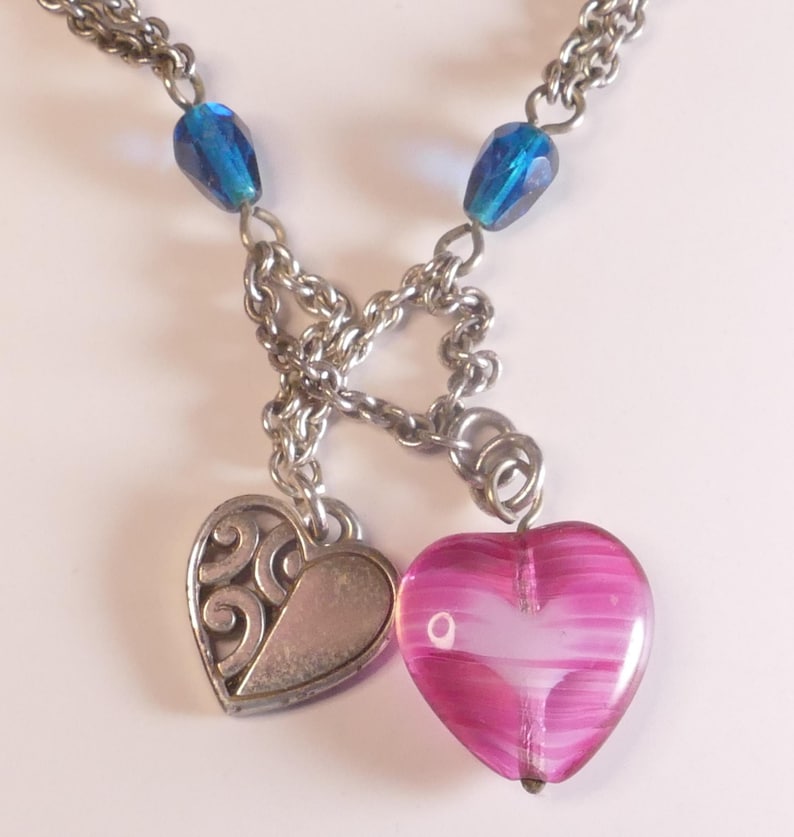 Puede incluir: Un collar de cadena de metal plateado con dos dijes en forma de coraz&oacute;n. Un dije es un coraz&oacute;n plateado s&oacute;lido con un patr&oacute;n de remolinos en relieve, y el otro es un coraz&oacute;n de vidrio rosa transl&uacute;cido con rayas blancas. Dos cuentas azules est&aacute;n unidas.
