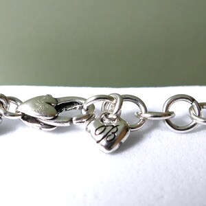 K&ouml;nnte beinhalten: Nahaufnahme eines silbernen Armbands mit herzf&ouml;rmigem Anh&auml;nger. Das Armband hat ein geflochtenes Design und einen Karabinerverschluss. Ein kleiner Herzanh&auml;nger mit dem Buchstaben "B" h&auml;ngt am Verschluss. Der Hintergrund ist ein sanftes Gr&uuml;n.