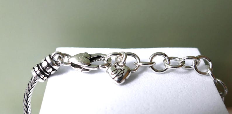 K&ouml;nnte beinhalten: Nahaufnahme eines silbernen Armbands mit herzf&ouml;rmigem Anh&auml;nger. Das Armband hat ein geflochtenes Design und einen Karabinerverschluss. Ein kleiner Herzanh&auml;nger mit dem Buchstaben "B" h&auml;ngt am Verschluss. Der Hintergrund ist ein sanftes Gr&uuml;n.