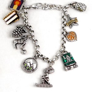 May include: Silver charm bracelet featuring various charms, including a pinecone, a mountain goat, a leaf, and a backpack. The bracelet also has a charm that says "Denver" and a skier. The bracelet is on a white background.