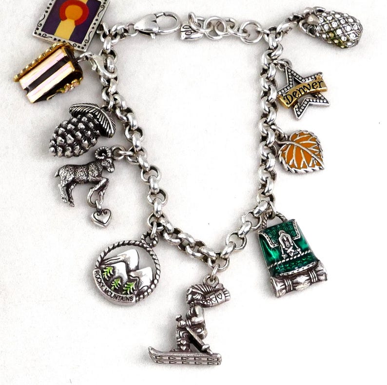 May include: Silver charm bracelet featuring various charms, including a pinecone, a mountain goat, a leaf, and a backpack. The bracelet also has a charm that says "Denver" and a skier. The bracelet is on a white background.