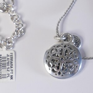 May include: A silver-toned necklace featuring a round pendant with an intricate cut-out filigree pattern and a matching chain. A white rectangular tag is attached, printed with FERRARA PT NK, JL4760 SZ: PL, 843036037325, and Made in TAIWAN.