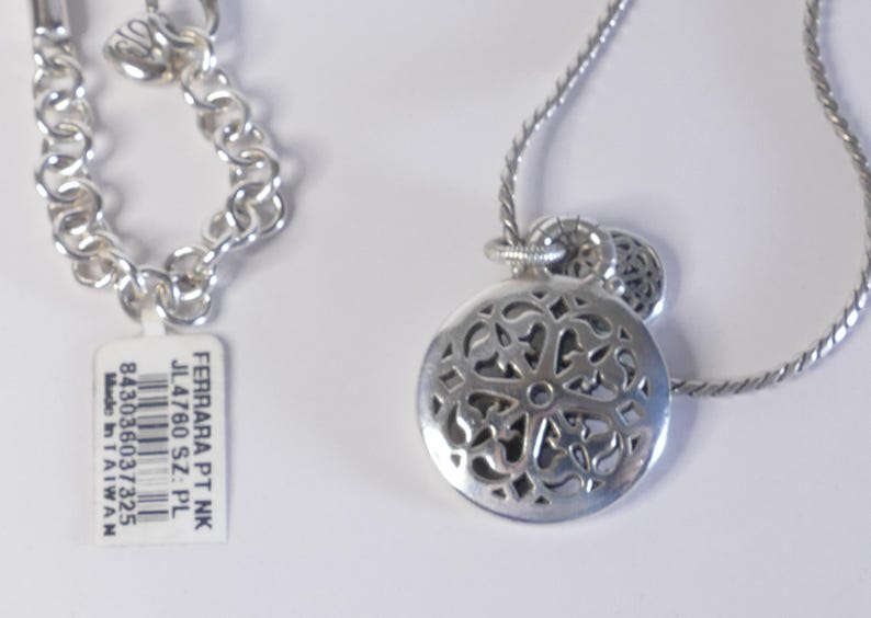 May include: A silver-toned necklace featuring a round pendant with an intricate cut-out filigree pattern and a matching chain. A white rectangular tag is attached, printed with FERRARA PT NK, JL4760 SZ: PL, 843036037325, and Made in TAIWAN.
