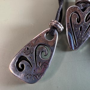 May include: Two silver-toned metal pendants with cut-out designs, each suspended on a black cord. The pendants feature engraved floral and swirling patterns, with a small metal bead accenting the cord above each piece.