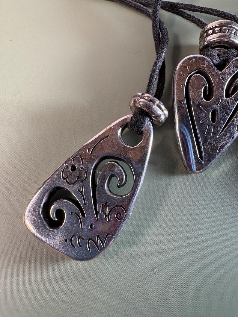 May include: Two silver-toned metal pendants with cut-out designs, each suspended on a black cord. The pendants feature engraved floral and swirling patterns, with a small metal bead accenting the cord above each piece.