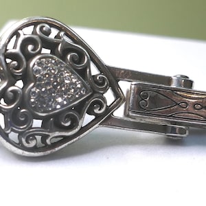 Puede incluir: Cierre plateado en forma de coraz&oacute;n con un dise&ntilde;o de filigrana y un coraz&oacute;n central incrustado con peque&ntilde;os cristales brillantes. El cierre tiene un estilo ornamentado y decorativo, con un cierre de bisagra y un patr&oacute;n grabado detallado.
