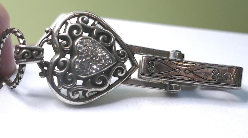 Puede incluir: Cierre plateado en forma de coraz&oacute;n con un dise&ntilde;o de filigrana y un coraz&oacute;n central incrustado con peque&ntilde;os cristales brillantes. El cierre tiene un estilo ornamentado y decorativo, con un cierre de bisagra y un patr&oacute;n grabado detallado.