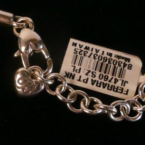 May include: A silver-toned metal chain necklace featuring a heart-shaped lobster clasp and a small heart charm with an engraved letter. A white rectangular tag is attached, displaying a barcode and the text FERRARA PT NK JL4760 SZ PL Made in TAIWAN.