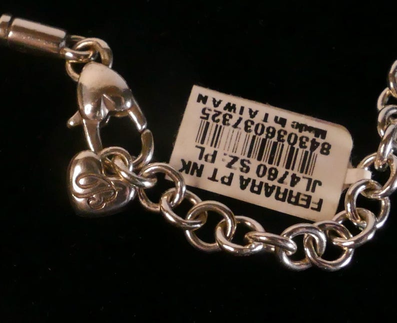 May include: A silver-toned metal chain necklace featuring a heart-shaped lobster clasp and a small heart charm with an engraved letter. A white rectangular tag is attached, displaying a barcode and the text FERRARA PT NK JL4760 SZ PL Made in TAIWAN.
