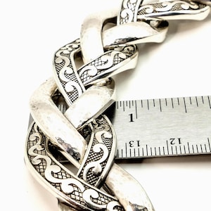 May include: Close-up of a silver-toned chain bracelet with an intricate, textured design. The bracelet's links are approximately 1 inch wide. The design features a hammered finish and detailed scrollwork, creating a bold, statement piece.