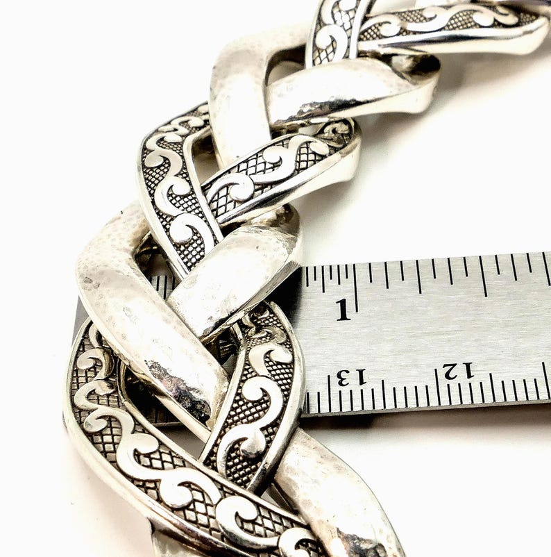 May include: Close-up of a silver-toned chain bracelet with an intricate, textured design. The bracelet's links are approximately 1 inch wide. The design features a hammered finish and detailed scrollwork, creating a bold, statement piece.