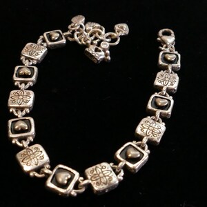 May include: Silver-toned bracelet with square links featuring heart and floral designs. The bracelet has a lobster clasp closure and a chain for adjusting the length. The bracelet is displayed on a black surface.