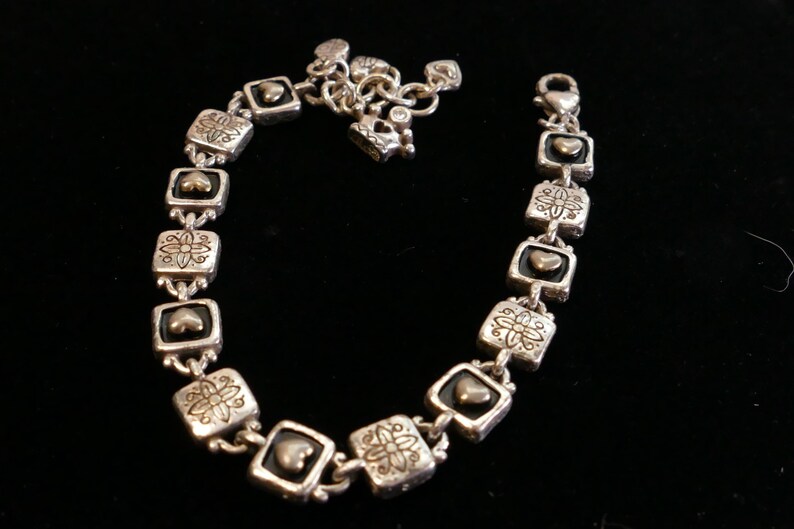May include: Silver-toned bracelet with square links featuring heart and floral designs. The bracelet has a lobster clasp closure and a chain for adjusting the length. The bracelet is displayed on a black surface.
