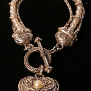 May include: Silver-toned bracelet featuring a heart-shaped pendant with a pearl accent. The bracelet has multiple strands of faux pearls and a decorative clasp. The heart pendant is detailed with floral designs.