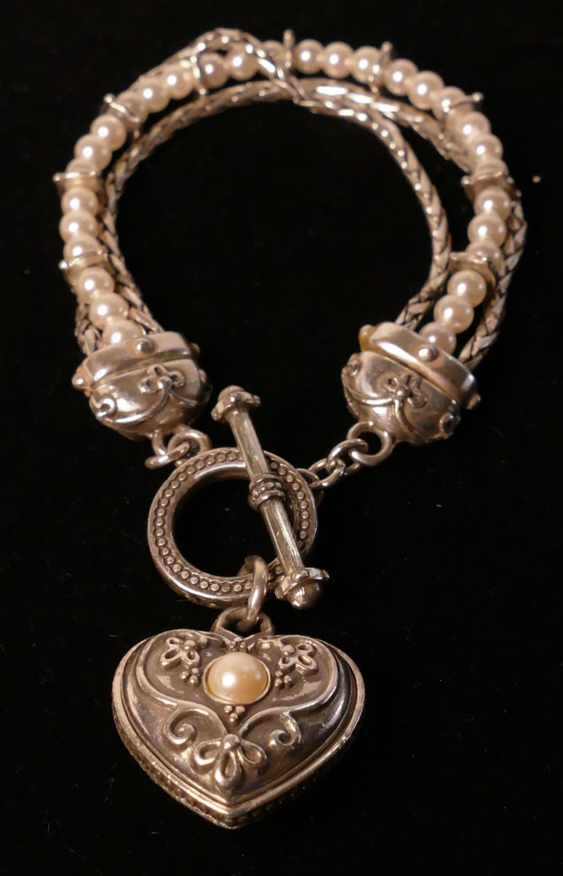 May include: Silver-toned bracelet featuring a heart-shaped pendant with a pearl accent. The bracelet has multiple strands of faux pearls and a decorative clasp. The heart pendant is detailed with floral designs.
