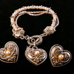 May include: A silver-toned jewelry set featuring a bracelet and two heart-shaped pieces. The bracelet has multiple strands with faux pearls and decorative clasps. The heart-shaped pieces have a central pearl and ornate detailing.