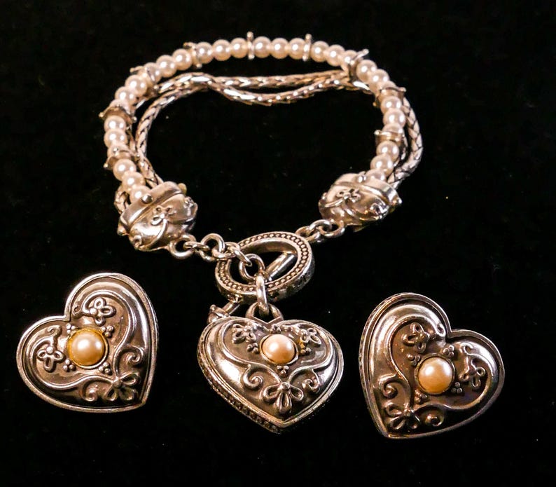 May include: A silver-toned jewelry set featuring a bracelet and two heart-shaped pieces. The bracelet has multiple strands with faux pearls and decorative clasps. The heart-shaped pieces have a central pearl and ornate detailing.