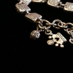 May include: Silver-toned charm bracelet with square links and decorative patterns. The bracelet features several charms, including a heart, a crown, and a small, round charm with a gemstone. The bracelet is set against a black background.