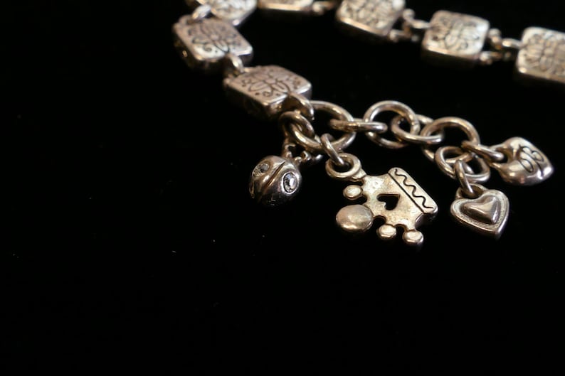 May include: Silver-toned charm bracelet with square links and decorative patterns. The bracelet features several charms, including a heart, a crown, and a small, round charm with a gemstone. The bracelet is set against a black background.