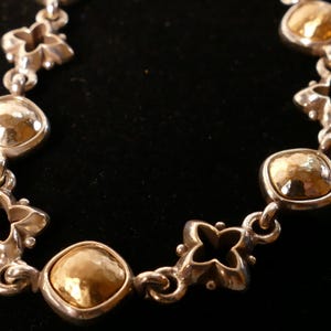 May include: A silver bracelet with gold-coloured, hammered-metal details. The bracelet has alternating quatrefoil and square-shaped links. The gold elements have a textured, dimpled appearance, set against a black background.