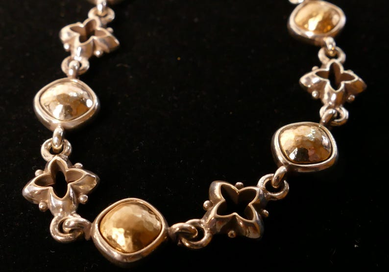 May include: A silver bracelet with gold-coloured, hammered-metal details. The bracelet has alternating quatrefoil and square-shaped links. The gold elements have a textured, dimpled appearance, set against a black background.