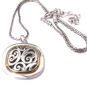 May include: Silver-tone necklace with a square pendant featuring a gold-tone inner frame and an openwork design. The chain is a braided style, and the pendant has a decorative, swirling pattern.