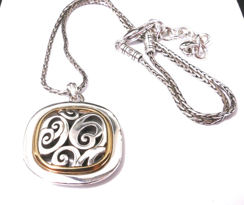 May include: Silver-tone necklace with a square pendant featuring a gold-tone inner frame and an openwork design. The chain is a braided style, and the pendant has a decorative, swirling pattern.