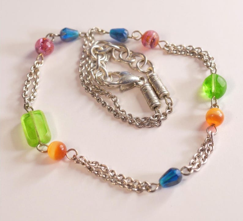 Puede incluir: Una pulsera de doble cadena en tono plateado con un surtido de cuentas de vidrio de colores. Las cuentas incluyen formas rectangulares de color verde transl&uacute;cido, esferas redondas naranjas y rosas, y cuentas azules en forma de l&aacute;grima, unidas por eslabones.