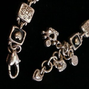 May include: Silver-toned bracelet with a variety of charms and decorative elements. The bracelet features square links with floral and heart designs, along with heart-shaped clasps and dangling charms. The background is black.