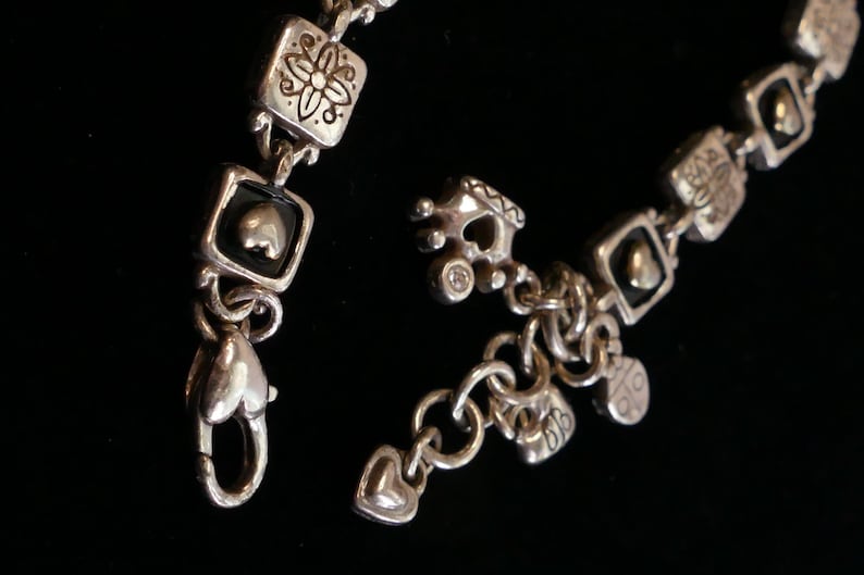 May include: Silver-toned bracelet with a variety of charms and decorative elements. The bracelet features square links with floral and heart designs, along with heart-shaped clasps and dangling charms. The background is black.
