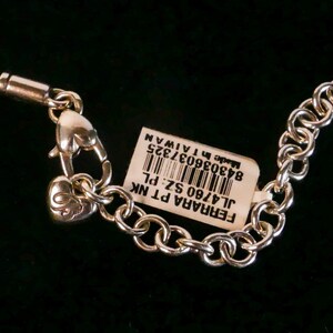 May include: A silver-toned metal chain necklace featuring a heart-shaped lobster clasp and a small heart charm with an engraved letter. A white rectangular product tag is attached, displaying a barcode and the text FERRARA PT NK JL4760 SZ: PL.