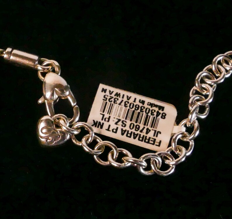 May include: A silver-toned metal chain necklace featuring a heart-shaped lobster clasp and a small heart charm with an engraved letter. A white rectangular product tag is attached, displaying a barcode and the text FERRARA PT NK JL4760 SZ: PL.