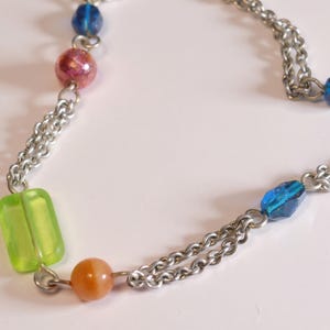 Puede incluir: Un collar de cadena de metal plateado con una variedad de cuentas de colores, que incluye una cuenta rectangular de color verde lima, una cuenta redonda naranja, una cuenta rosa moteada y varias cuentas azules facetadas entre los eslabones.