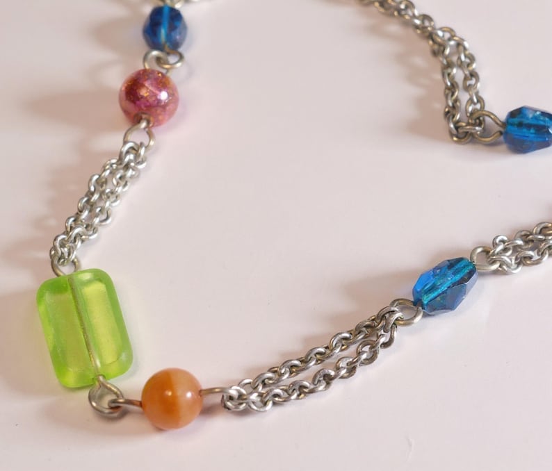 Puede incluir: Un collar de cadena de metal plateado con una variedad de cuentas de colores, que incluye una cuenta rectangular de color verde lima, una cuenta redonda naranja, una cuenta rosa moteada y varias cuentas azules facetadas entre los eslabones.