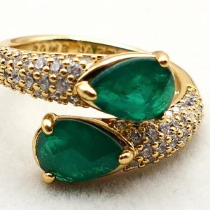 May include: A gold ring featuring two pear-shaped, vibrant green emeralds. The ring's band is encrusted with small, sparkling white gemstones. The design is elegant and eye-catching, showcasing a luxurious jewelry piece.