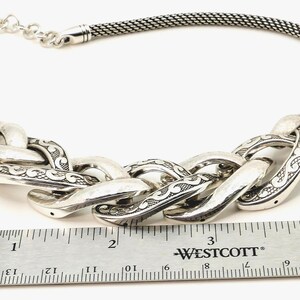 May include: Silver-toned necklace featuring a chunky chain design with etched detailing. The necklace includes a mesh chain and a series of large, interlocking links. A ruler is placed next to the necklace for scale.