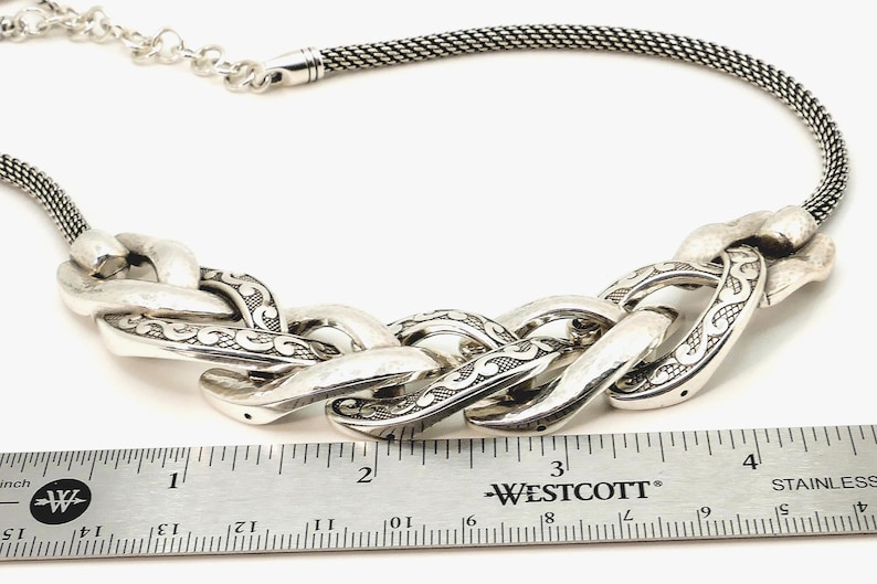 May include: Silver-toned necklace featuring a chunky chain design with etched detailing. The necklace includes a mesh chain and a series of large, interlocking links. A ruler is placed next to the necklace for scale.