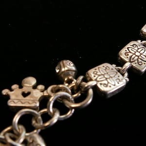May include: Silver-toned bracelet with decorative charms. The bracelet features square links with floral designs, a crown charm with a heart cutout, and a small, gem-studded cube charm. The background is black.