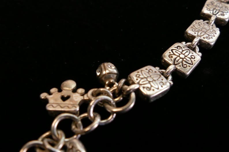 May include: Silver-toned bracelet with decorative charms. The bracelet features square links with floral designs, a crown charm with a heart cutout, and a small, gem-studded cube charm. The background is black.