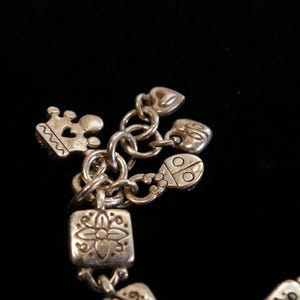May include: Silver charm bracelet with various charms, including a crown, hearts, a ladybug, and a square charm with a floral design. The bracelet is made of linked chains and charms.