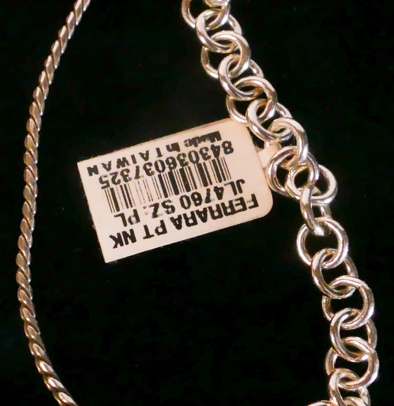 May include: A silver-toned necklace featuring a twisted chain segment and a series of circular links. A rectangular white tag is attached, displaying a barcode and the text: FERRARA PT NK, JL4760 SZ: PL, 843036037325, Made in TAIWAN.