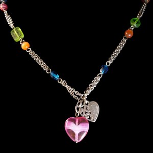 Puede incluir: Un collar de cadena de tono plateado con una variedad de cuentas coloridas que incluyen verde, naranja y azul. El collar tiene dos colgantes en forma de coraz&oacute;n que cuelgan del centro, uno de vidrio rosa transl&uacute;cido y otro de metal plateado.