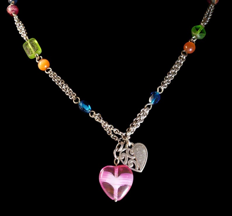 Puede incluir: Un collar de cadena de tono plateado con una variedad de cuentas coloridas que incluyen verde, naranja y azul. El collar tiene dos colgantes en forma de coraz&oacute;n que cuelgan del centro, uno de vidrio rosa transl&uacute;cido y otro de metal plateado.