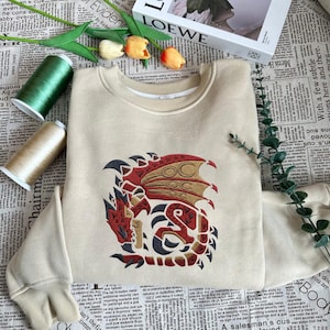 May include: A beige sweatshirt featuring a detailed embroidered dragon design in red, gold, and black. The dragon is centered on the front of the crew neck sweatshirt. The image also includes spools of thread and decorative flowers.