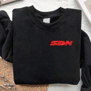 Könnte beinhalten: Schwarzes Sweatshirt mit Rundhalsausschnitt und roten, aufgestickten Buchstaben "SDN" auf der Brust. Das Sweatshirt hat einen gerippten Ausschnitt und Bündchen. Der Stoff scheint weich und bequem zu sein.
